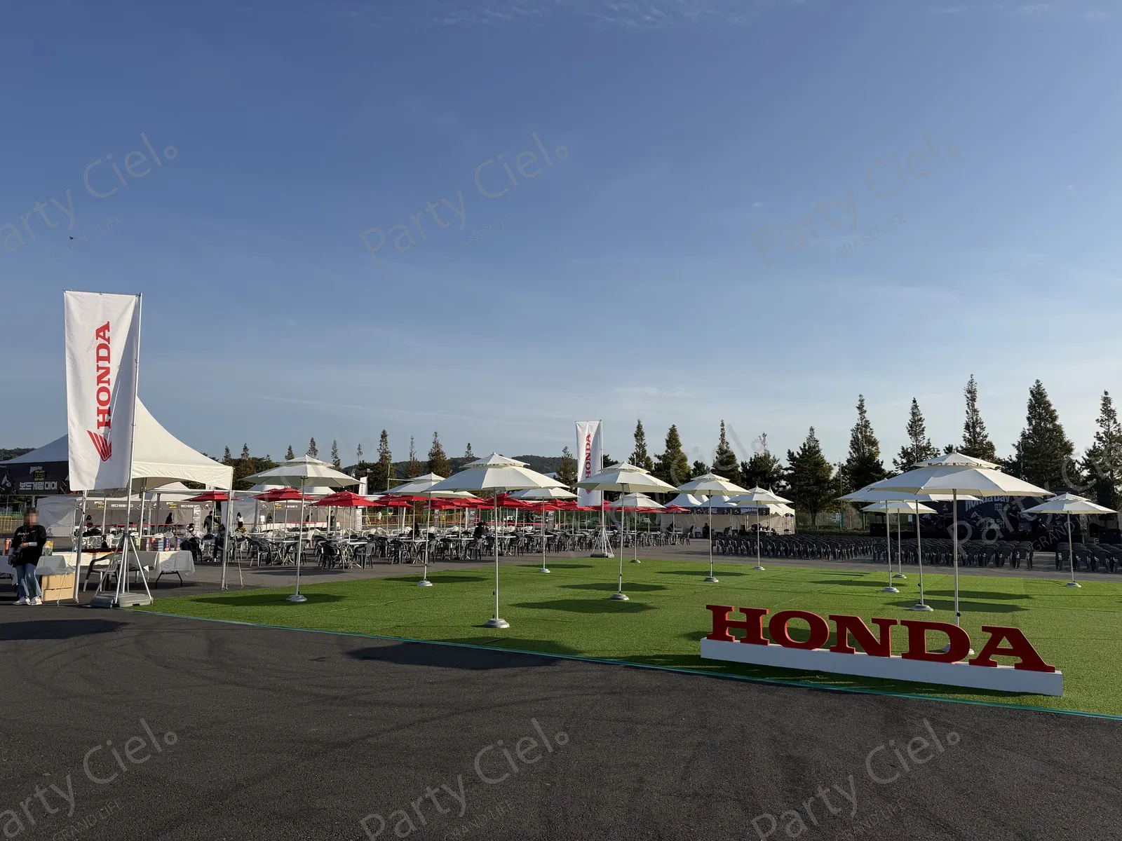 제11회 혼다데이 행사장 전경 — 인조잔디 위 빨간 입체 HONDA 조형물과 윙 로고 세로 깃발, 파라솔 아래 테이블과 의자가 늘어서 있고 멀리 HONDA DAY 백월과 흰 텐트, 야산이 보인다