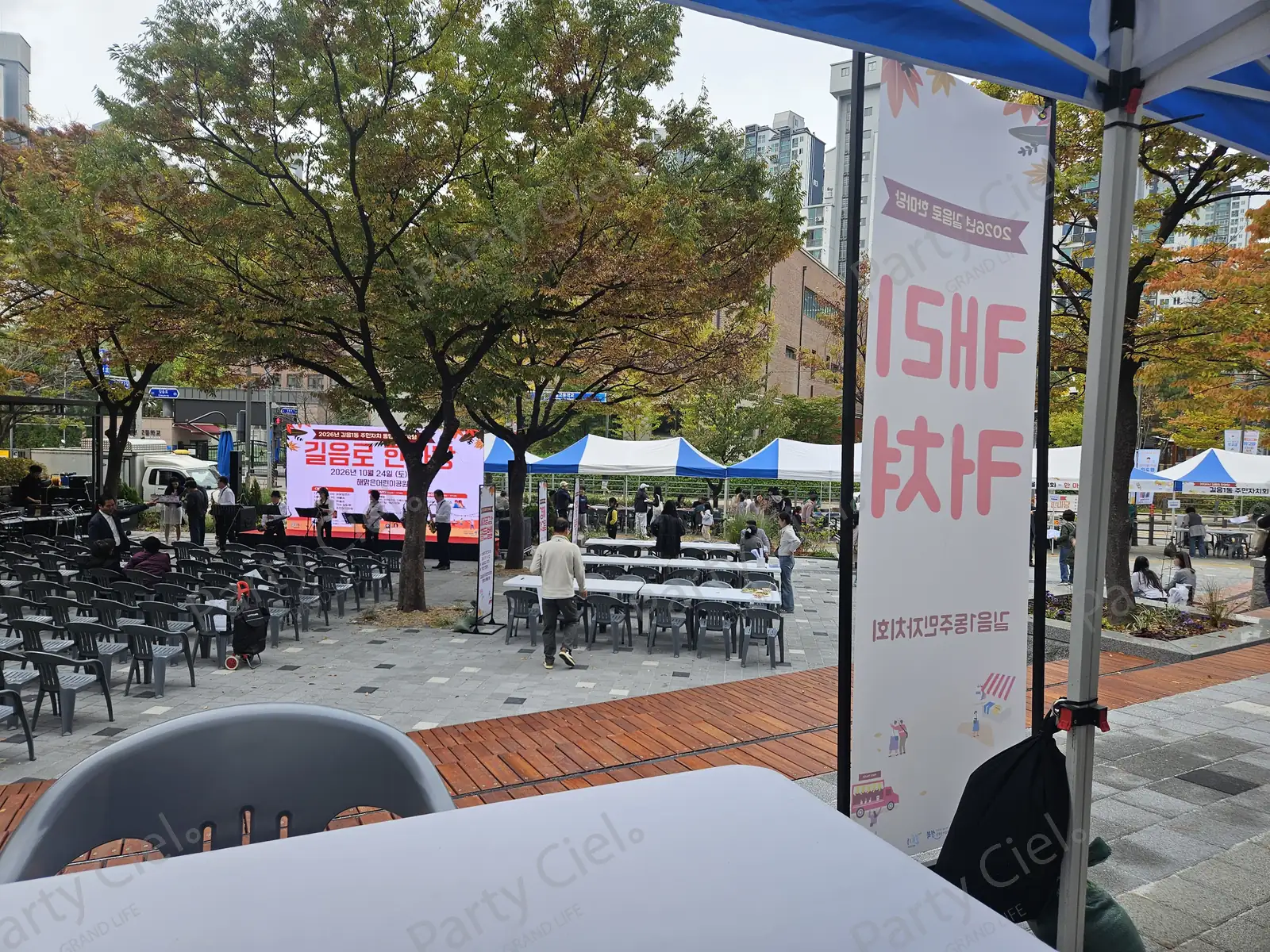 캐리커처 부스 그늘막 아래에서 바라본 공원 축제 행사장. 흰 테이블과 회색 의자 너머로 무대와 객석, 천막 부스가 보인다