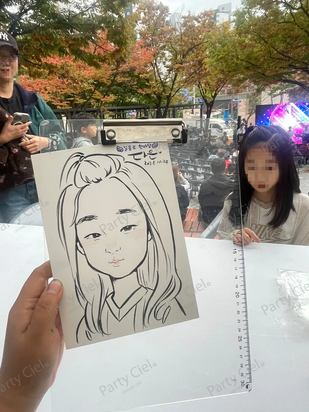 클립보드에 물린 종이에 긴 생머리 아이를 검은 펜선으로 그린 스피드 캐리커처. 볼과 입술에 옅은 분홍빛이 있고, 뒤로 그림의 주인공 아이와 객석이 보인다