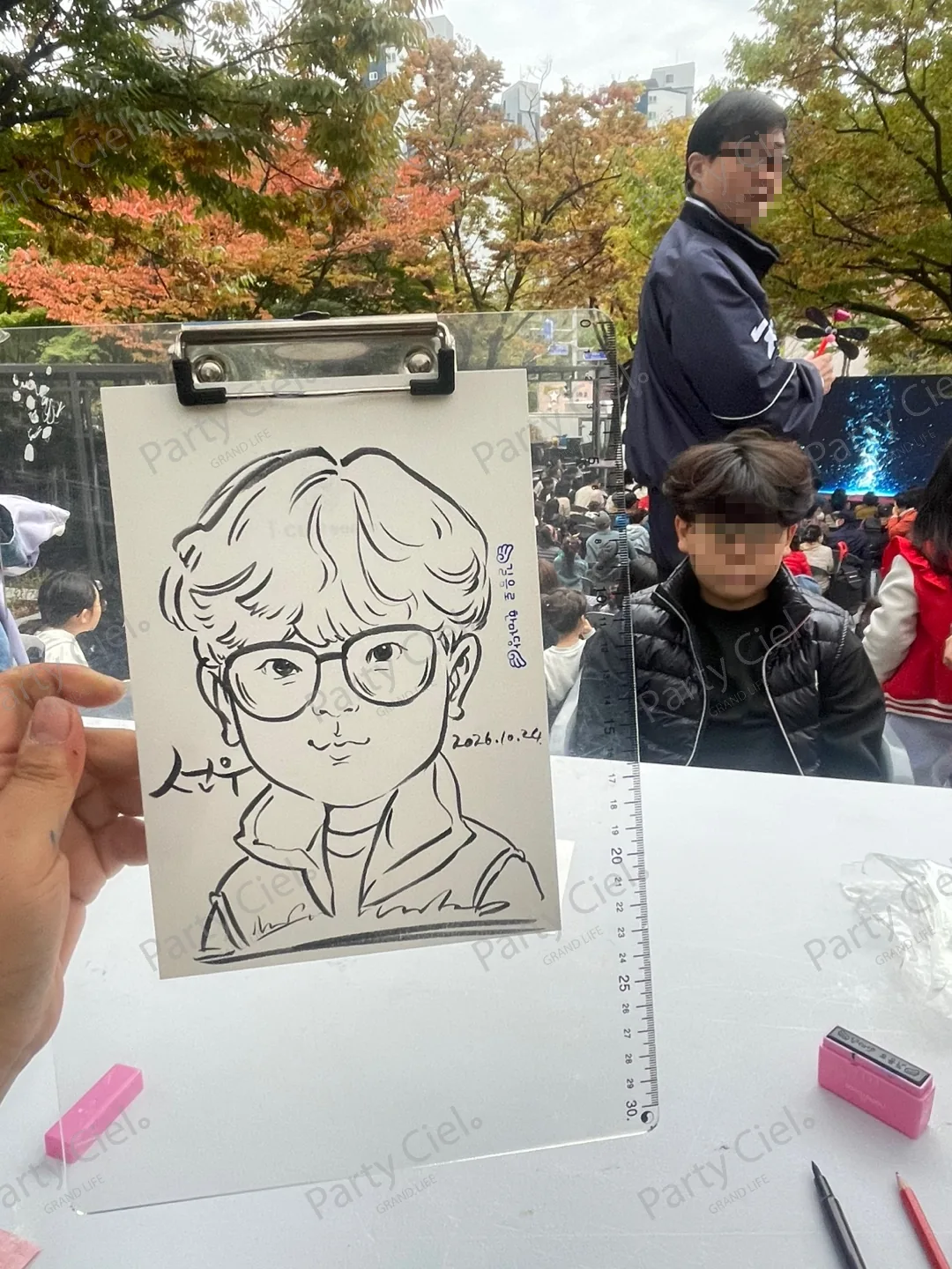 안경을 쓴 아이를 그린 스피드 캐리커처를 클립보드째 들어 보이는 모습. 테이블 위에 펜이 놓여 있다