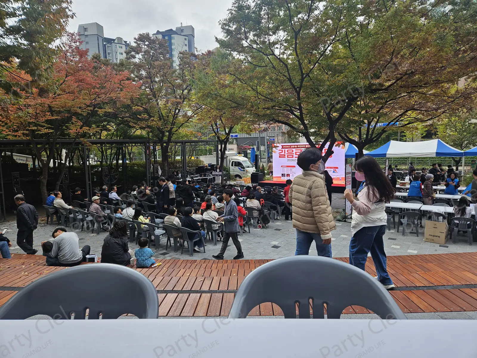 단풍 든 공원 축제 행사장. 무대 앞 객석에 주민들이 앉아 있고 오른쪽으로 파란 줄무늬 천막 부스가 늘어서 있다
