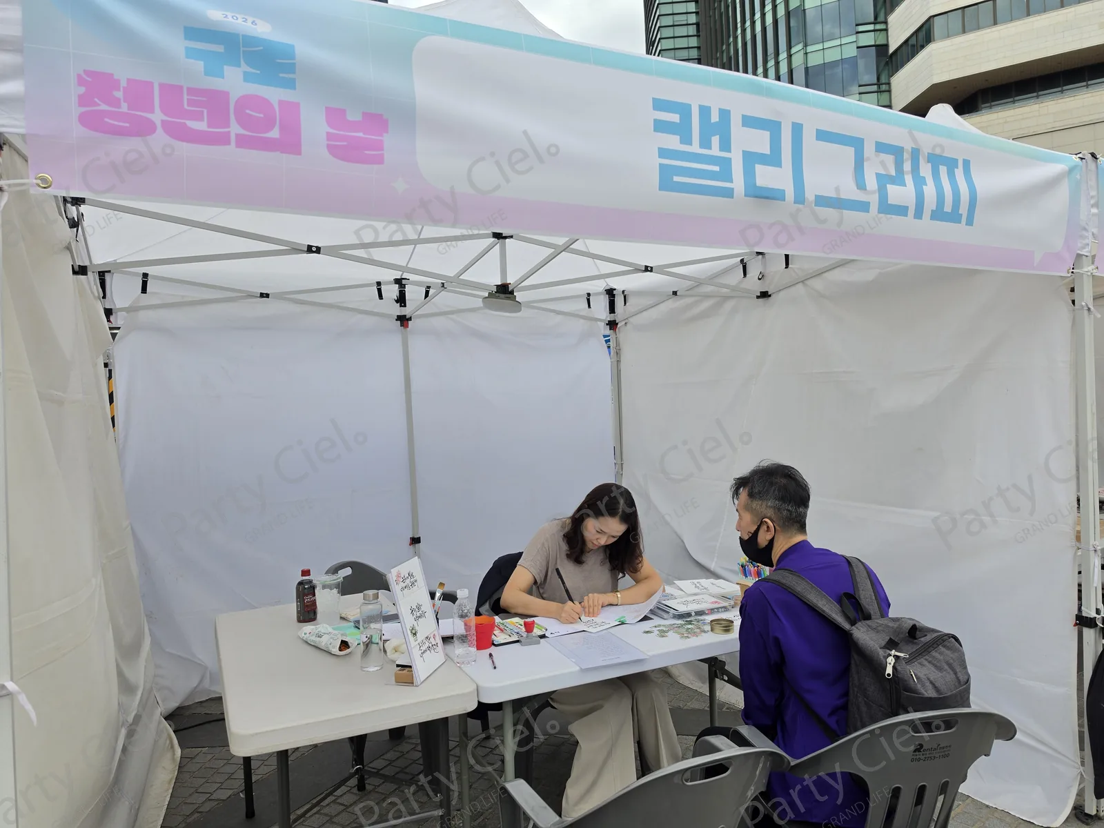 「2026 구로 청년의 날 · 캘리그라피」 현수막이 걸린 천막 부스에서 전문가가 종이에 문구를 쓰고 있는 모습