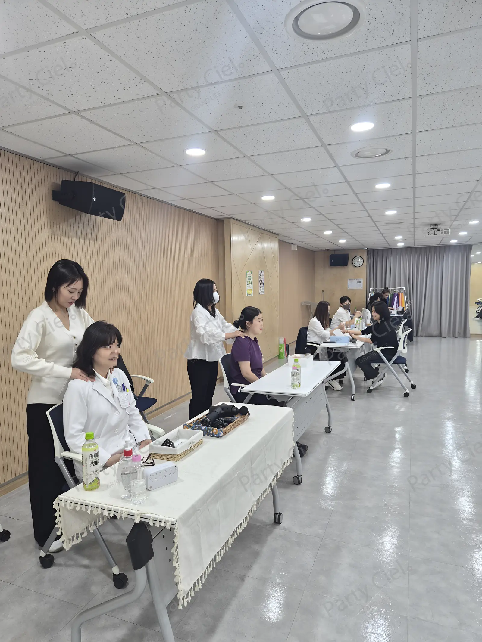 병원 실내 홀에 어깨마사지 두 자리와 핸드마사지 테이블이 나란히 놓이고 안쪽에 퍼스널컬러 부스가 보이는 행사 현장