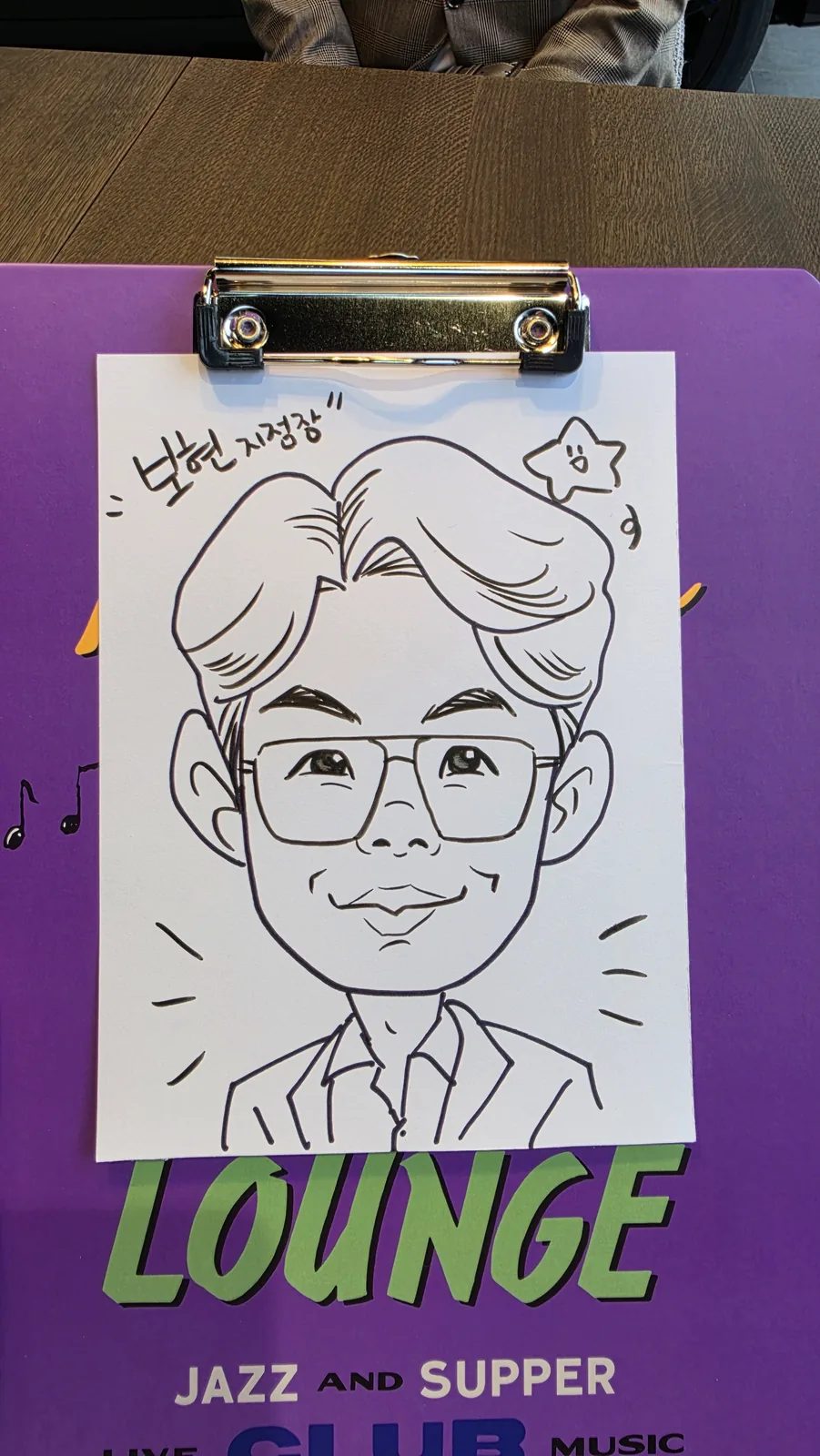 클립보드에 물린 흰 종이에 그려진 흑백 펜선 캐리커처. 안경을 쓰고 정장을 입은 성인 남성이 웃고 있는 상반신 그림으로, 주변에 집중선과 별 장식이 함께 그려져 있다.