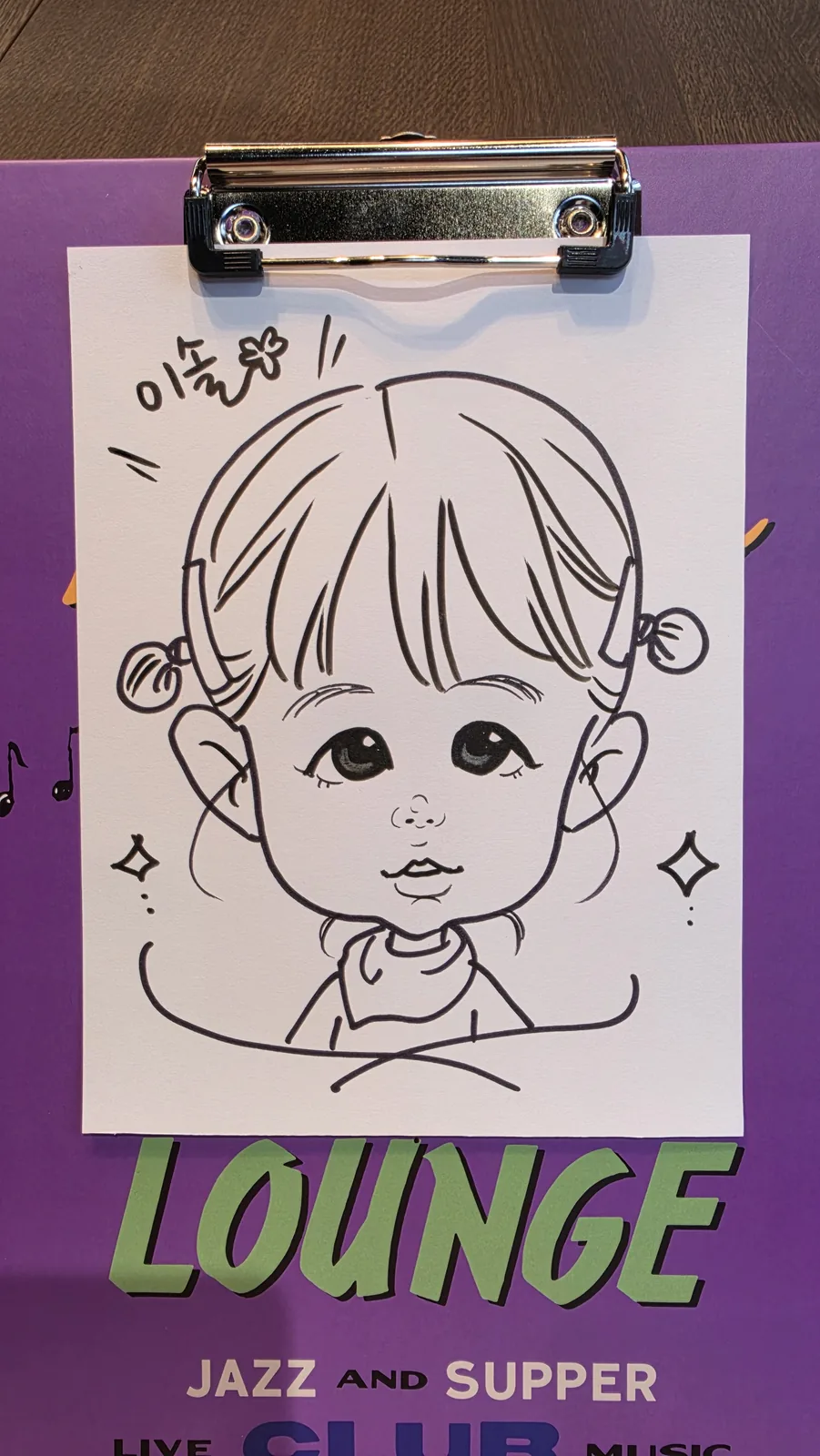 클립보드에 물린 흰 종이에 그려진 흑백 펜선 캐리커처. 양갈래로 머리를 묶고 턱받이를 한 아기가 큰 눈으로 정면을 보고 있으며, 양옆에 반짝임 장식이 그려져 있다.