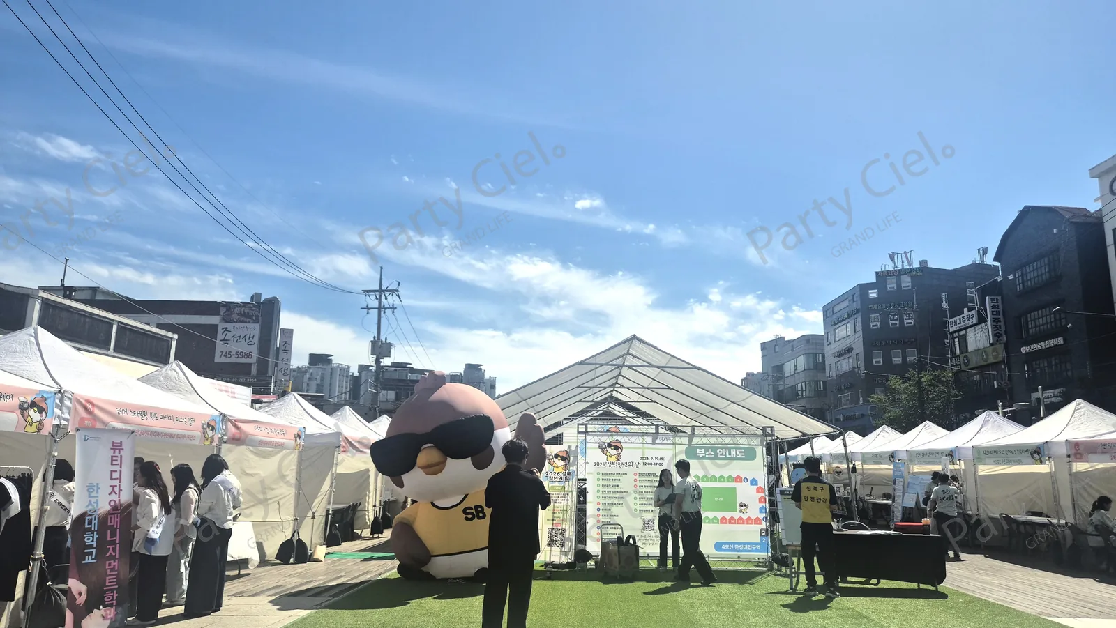 흰 천막 부스가 양옆으로 줄지어 선 광장 통로와 가운데 놓인 대형 캐릭터 조형물, 그 뒤 부스 안내도 입간판