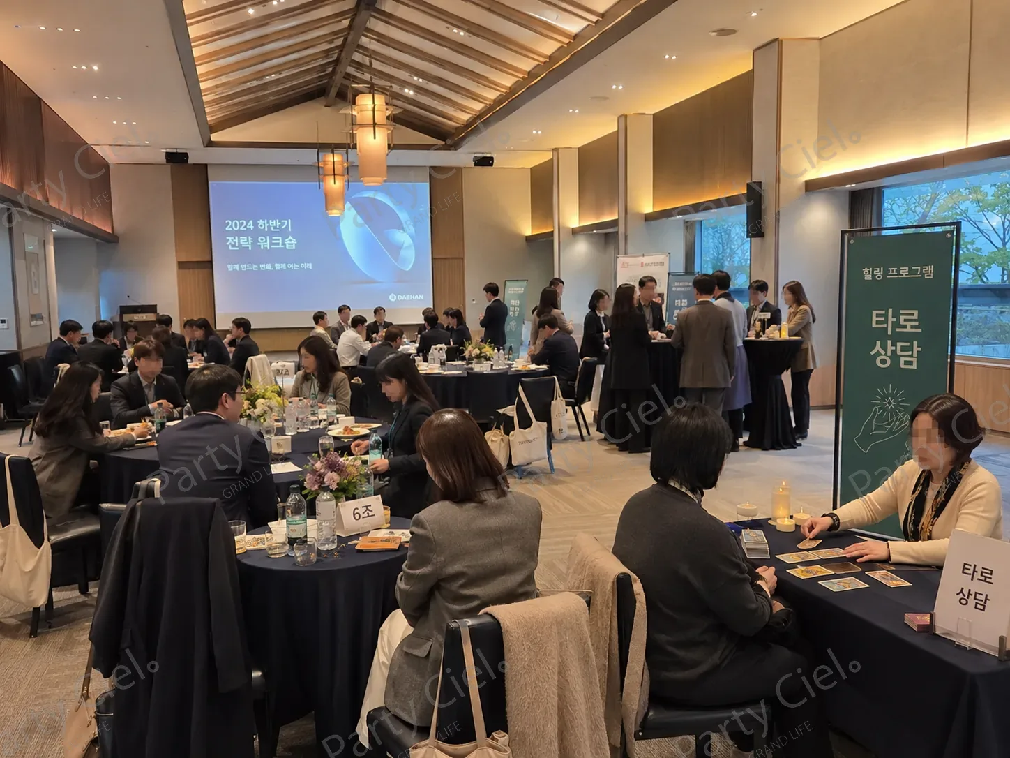 사내 하반기 전략 워크숍이 열린 연회장 — 조 번호 팻말이 놓인 원탁들 한쪽 끝 벽 가까이에 타로 상담 테이블과 힐링 프로그램 타로 상담이라고 적힌 세로 배너가 서 있다