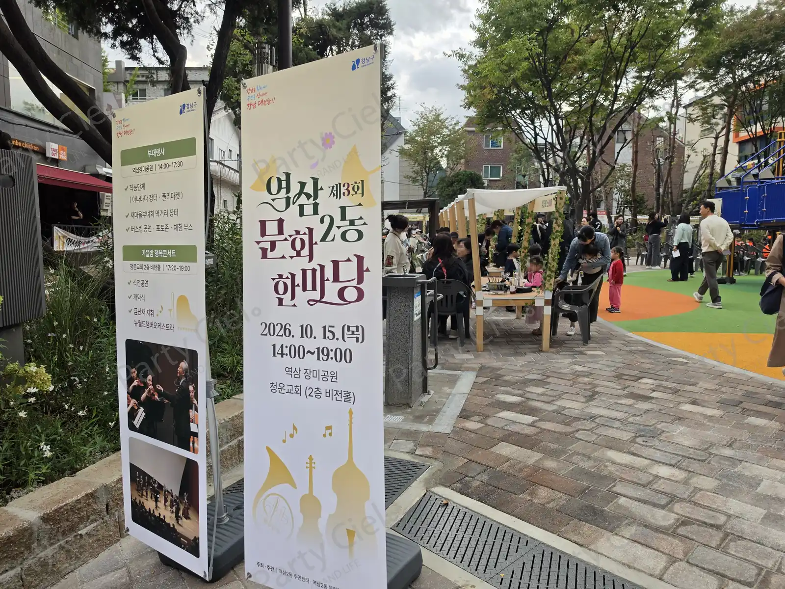 제3회 역삼2동문화한마당 입구에 세워진 행사 안내 배너 두 개와 공원 축제장 전경 — 오른쪽으로 체험부스 천막과 놀이터가 보인다