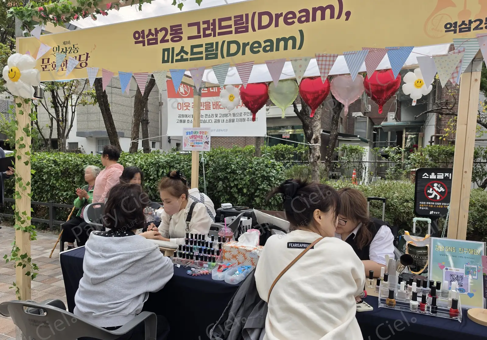 파고라 아래 두 자리에서 네일아트가 동시에 진행되는 모습 — 전문가 두 분이 각각 참가자의 손을 맡고 있고, 옆에 폴리시 진열대와 브러시통, 안내판이 놓여 있다