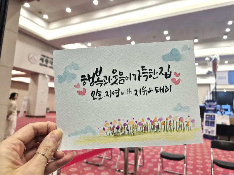 구름과 꽃밭 그림을 곁들인 가족 문구 캘리그라피 작품 카드 — 행사 현장 제작